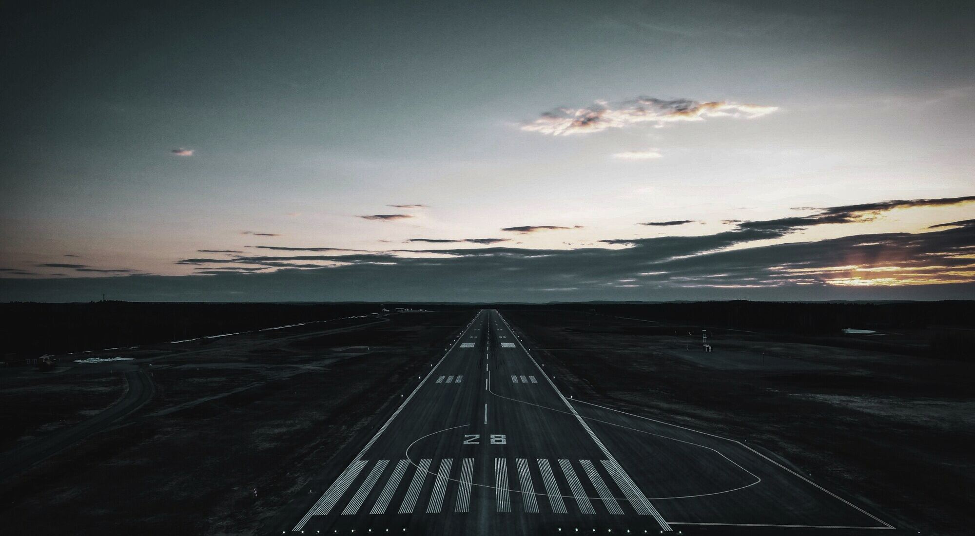 An empty airport runway at dusk, its markings running towards the horizon