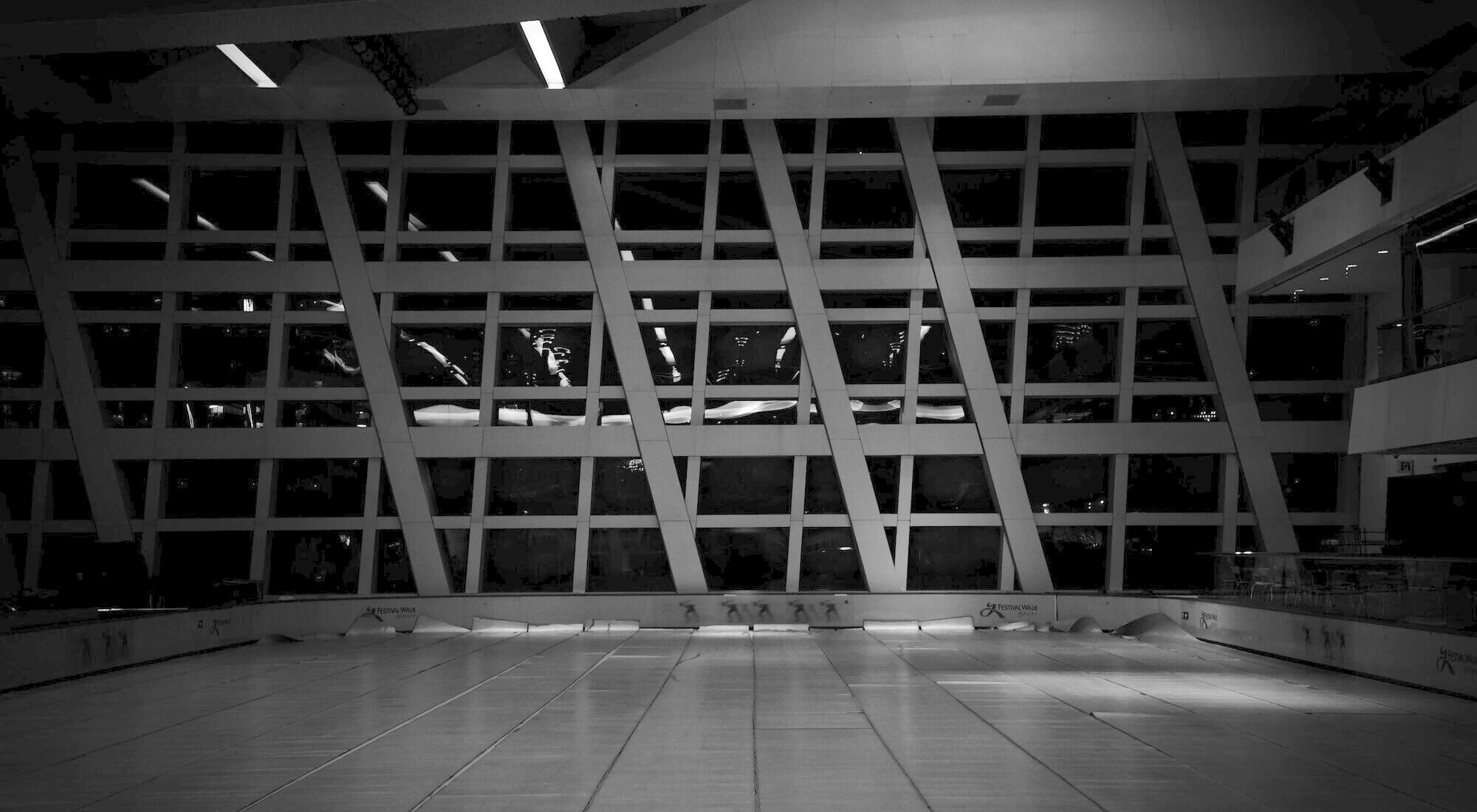 An empty public concourse at night, a structural glass wall above a reflective floor