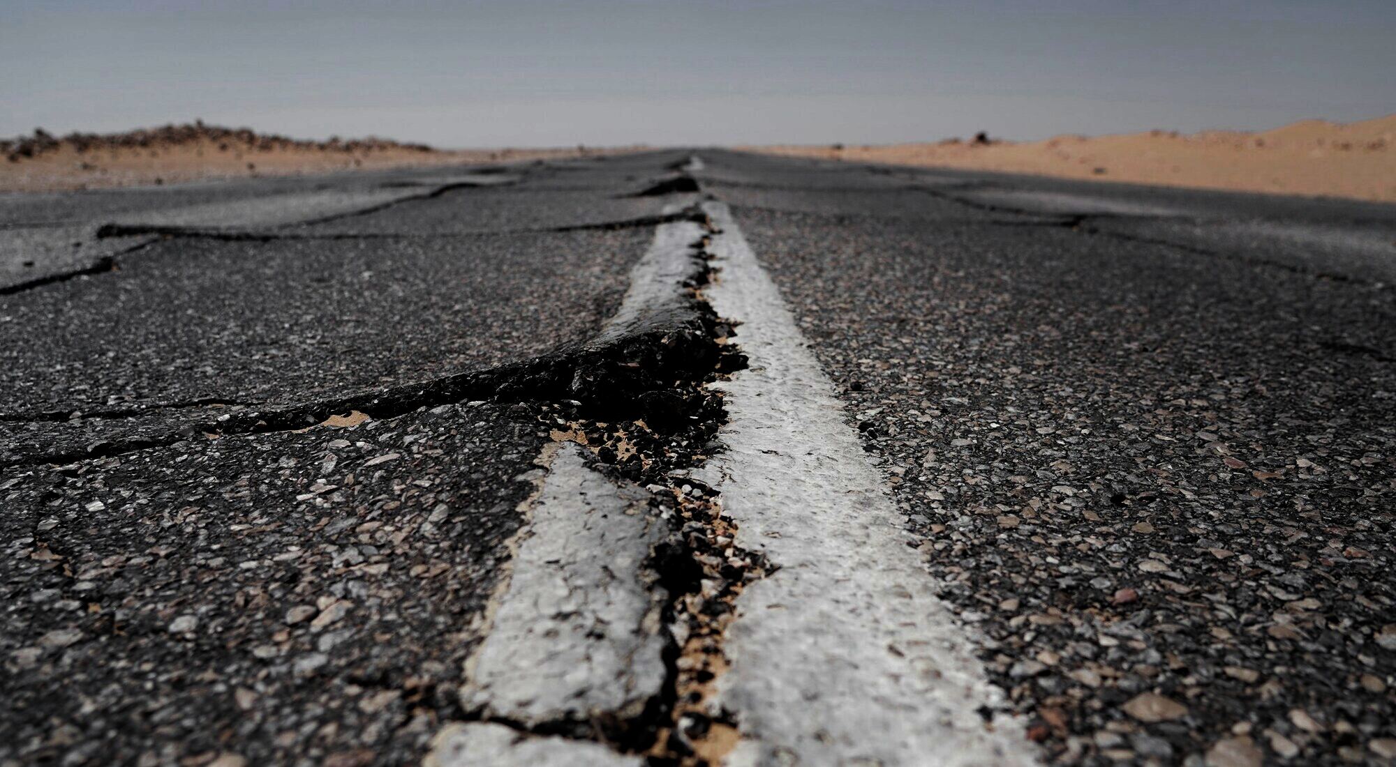 A cracked and broken public road, its centre line split, running into a dry landscape